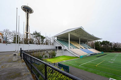 mutilevel grandstands with an angled roof - part of an athletic field below it and a wall behind it. The Space Needle is in  the background behind the wall