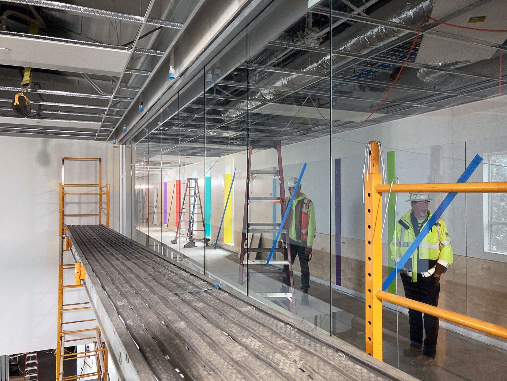 a scaffolding is in front of a partially installed glass wall. On th eother side of the glass wall, people are standing in a hallway