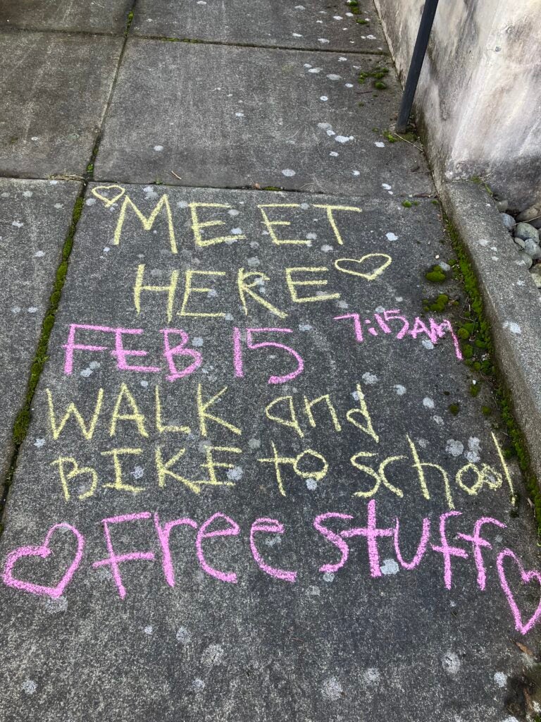 Sidewalk chalk that says "Meet here; Feb 15, Walk and bike to school; Free stuff"