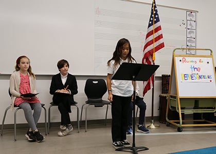 A student with two students behind them stand in front of a classroom at a music stand 