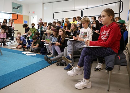 A group of students listens in a classroom to another student giving a speech