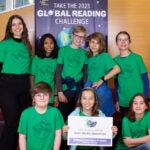 A group of students and a teacher gather for a photo in front of the library Global Reading Challenge banner.