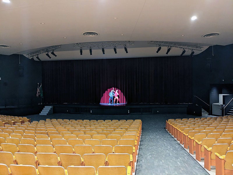 an auditorium with light brown seats looks toward a stage where two people are in the spotlight