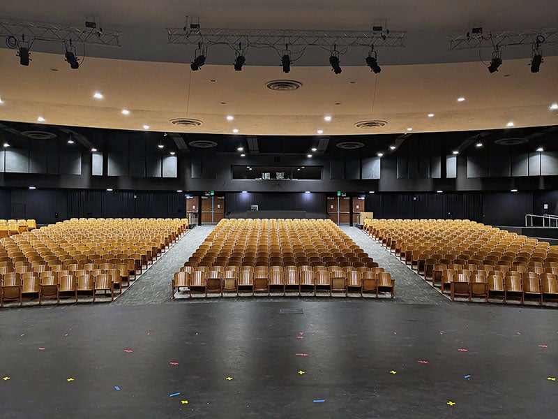 a view out into a auditorium with rows of light colored seats with dark walls, doors, and a control booth on the back wall