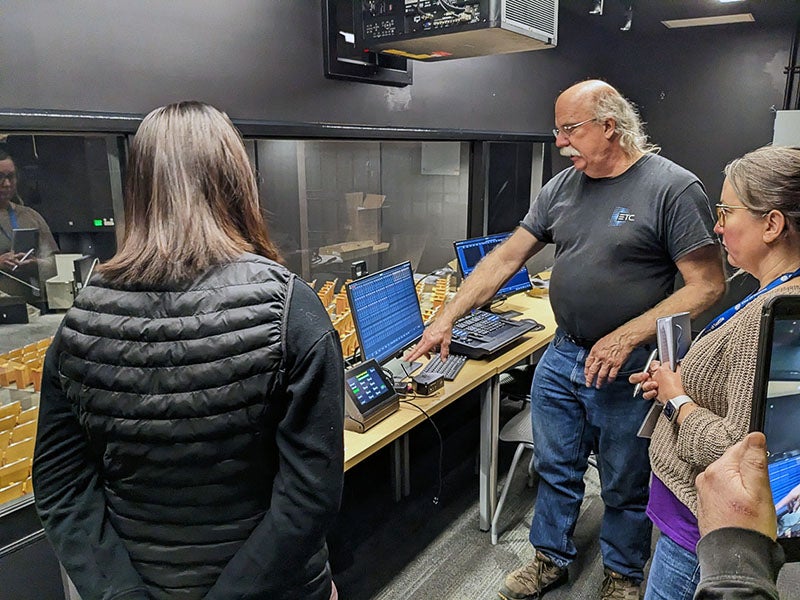 three people stand in a control booth with computers - a window looks out to auditorium seating below