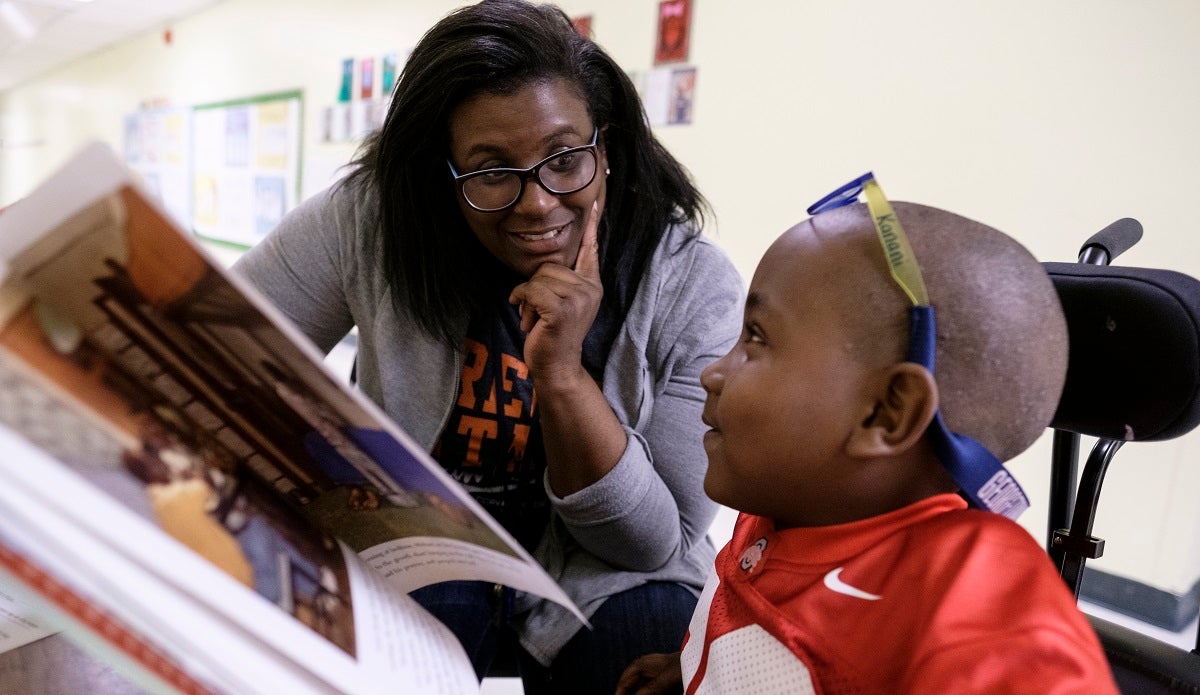 An educator and a young student talk together in a classroom while looking at a book.