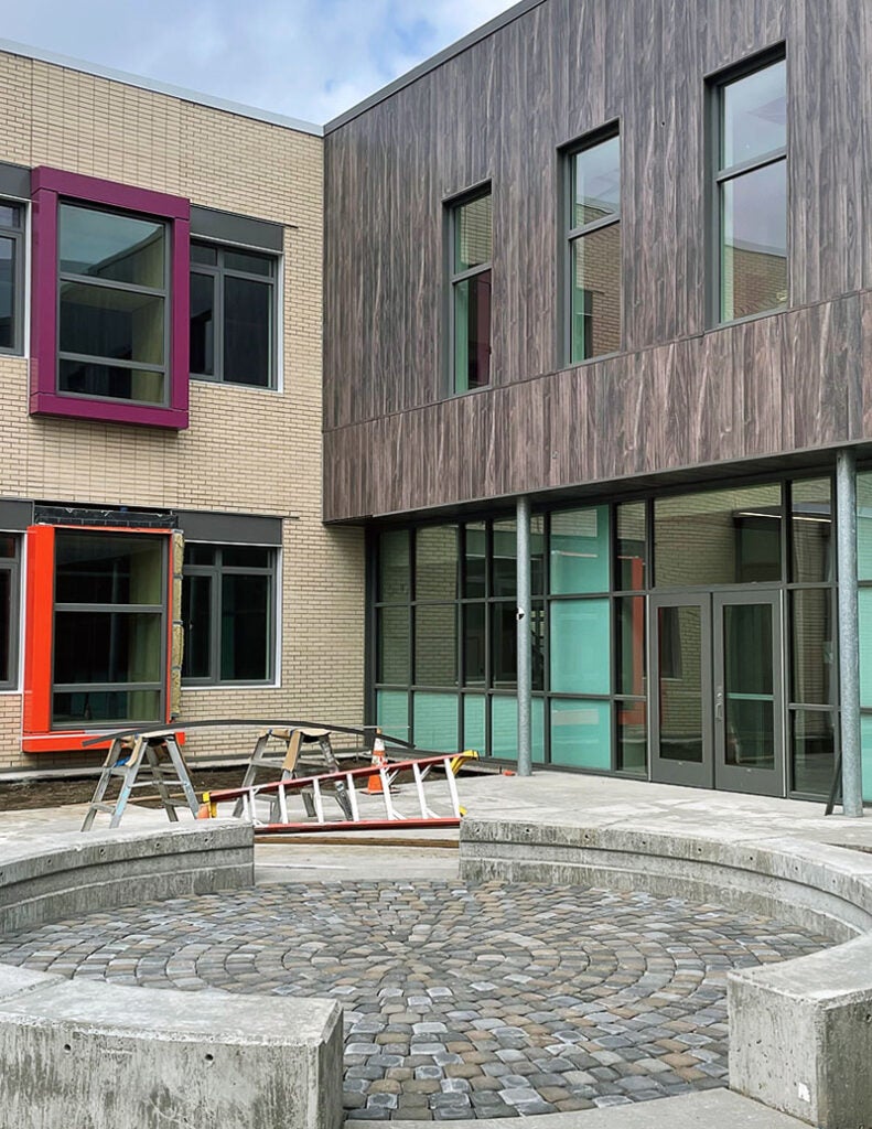 a round cobblestone patio is surrounded by two arcs that form a circle with space between them. The building behind forms a corner with one wall light brick with bright window frames and one wall of dark brown with doors and windows