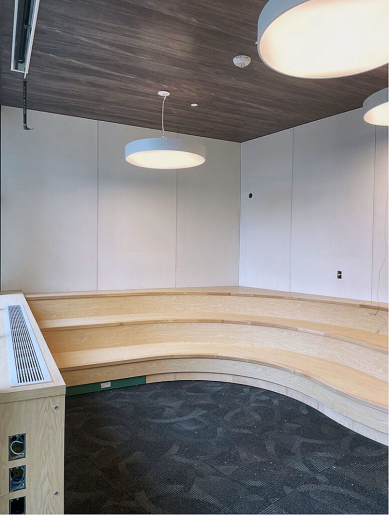 an interior corner has carpet, three tiers of curved benches, white wall panels, and a brown ceiling with circular lights hanging down