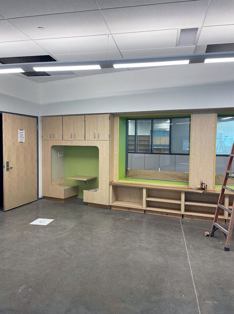 a classrom wall section has a nook below cabinets that includes two facing benches with a ledge between them. next to them is open shelving below a window. Both areas are highlighted by bright green accents