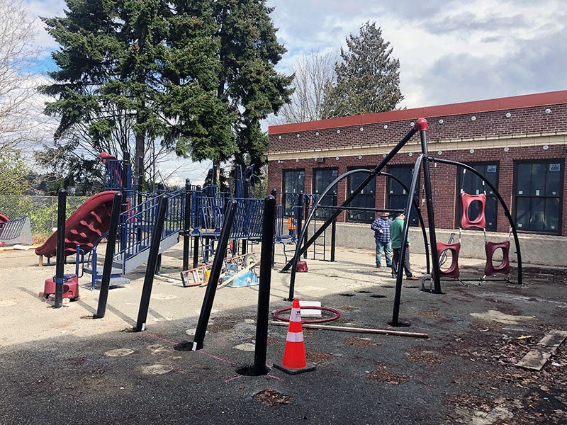 outside of a 1-story brick building, playground equipment is being installed. there are trees in the background