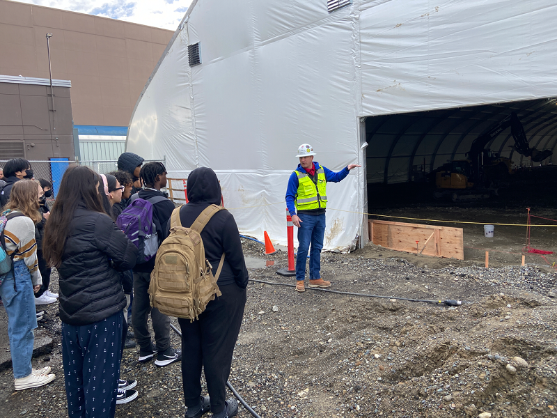 a group of people are watching while a person in a bright yellow vest and construction hat points at the opening to a large white tent. the ground is dirt.