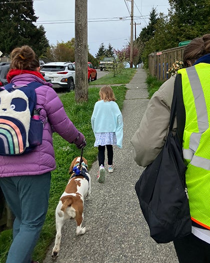 A student and two adults and one dog walk on a sidewalk.