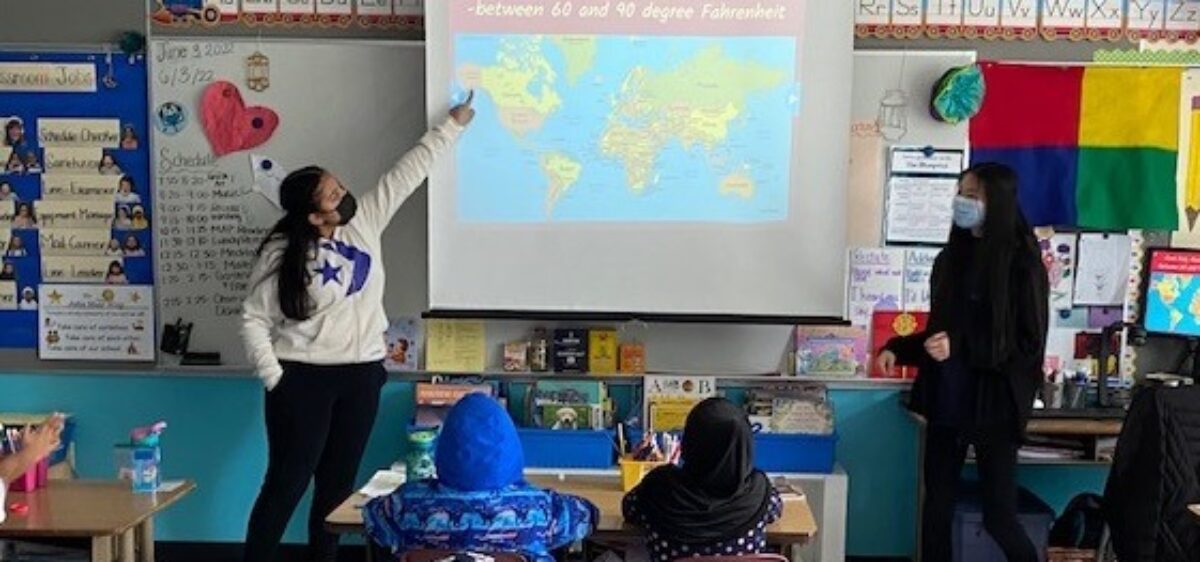 Two student teachers stand before elementary age students at a map of the world