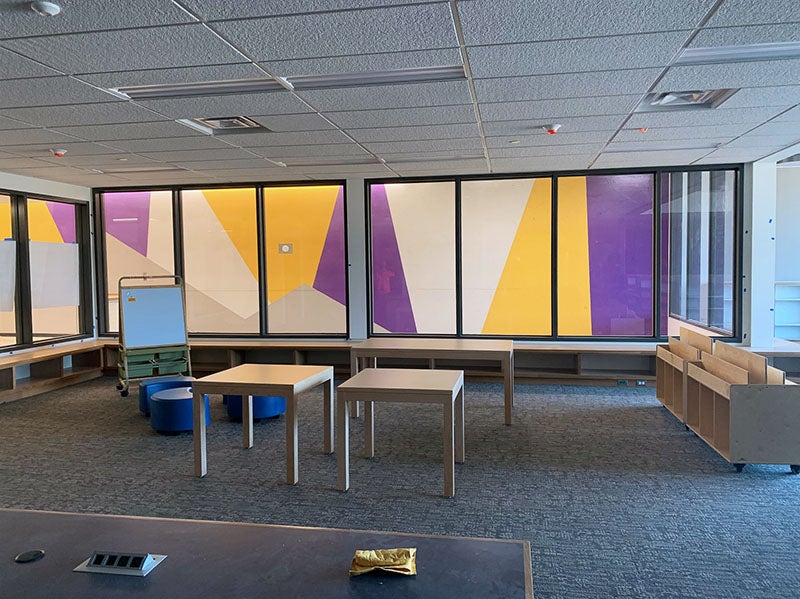 a wall of windows shows a bright colored mural on the wall outside. tables and book cases are in front of the windows