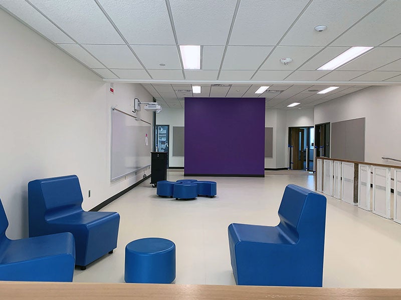 a group area has seating clusters, a learning wall, and a teacher station with a purple wall separating it from the hallway