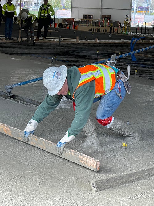 a person in boots with plastic wrapped at the top is wearing a safety vest and hard hat while wading in wet concrete and bending to smooth it with a piece of wood