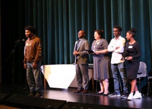 A group of students stands on a school stage during the award ceremony. One student is at a microphone.