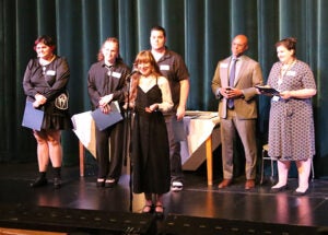 A group of students stands on a school stage during the award ceremony. One student is at a microphone.