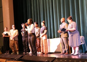 A group of students stands on a school stage during the award ceremony. One student is at a microphone.