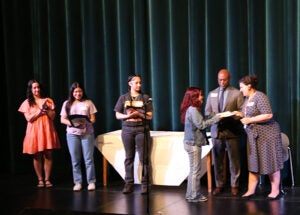 A group of students stands on a school stage during the award ceremony. One student accepts the award from School Board President Rankin