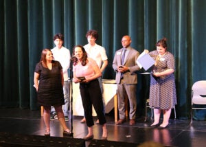 A group of students stands on a school stage during the award ceremony. One school counselor is at a microphone.