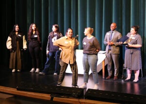 A group of students stands on a school stage during the award ceremony. Two school counselors are at a microphone.
