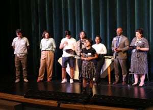 A group of students stands on a school stage during the award ceremony. One student is at a microphone.