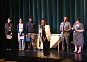 A group of students stands on a school stage during the award ceremony. One student is at a microphone.
