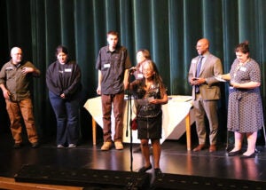 A group of students stands on a school stage during the award ceremony. One teacher is at a microphone.