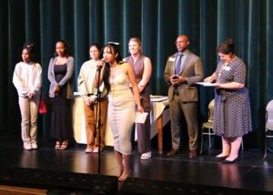 A group of students stands on a school stage during the award ceremony. One student is at a microphone.