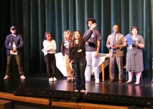 A group of students stands on a school stage during the award ceremony. One student is at a microphone.