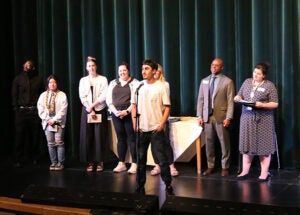 A group of students stands on a school stage during the award ceremony. One student is at a microphone.