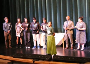 A group of students stands on a school stage during the award ceremony. One student is at a microphone.