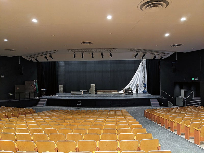 a view from behind theater seating toward a stage with a white curtain being installed at the the top