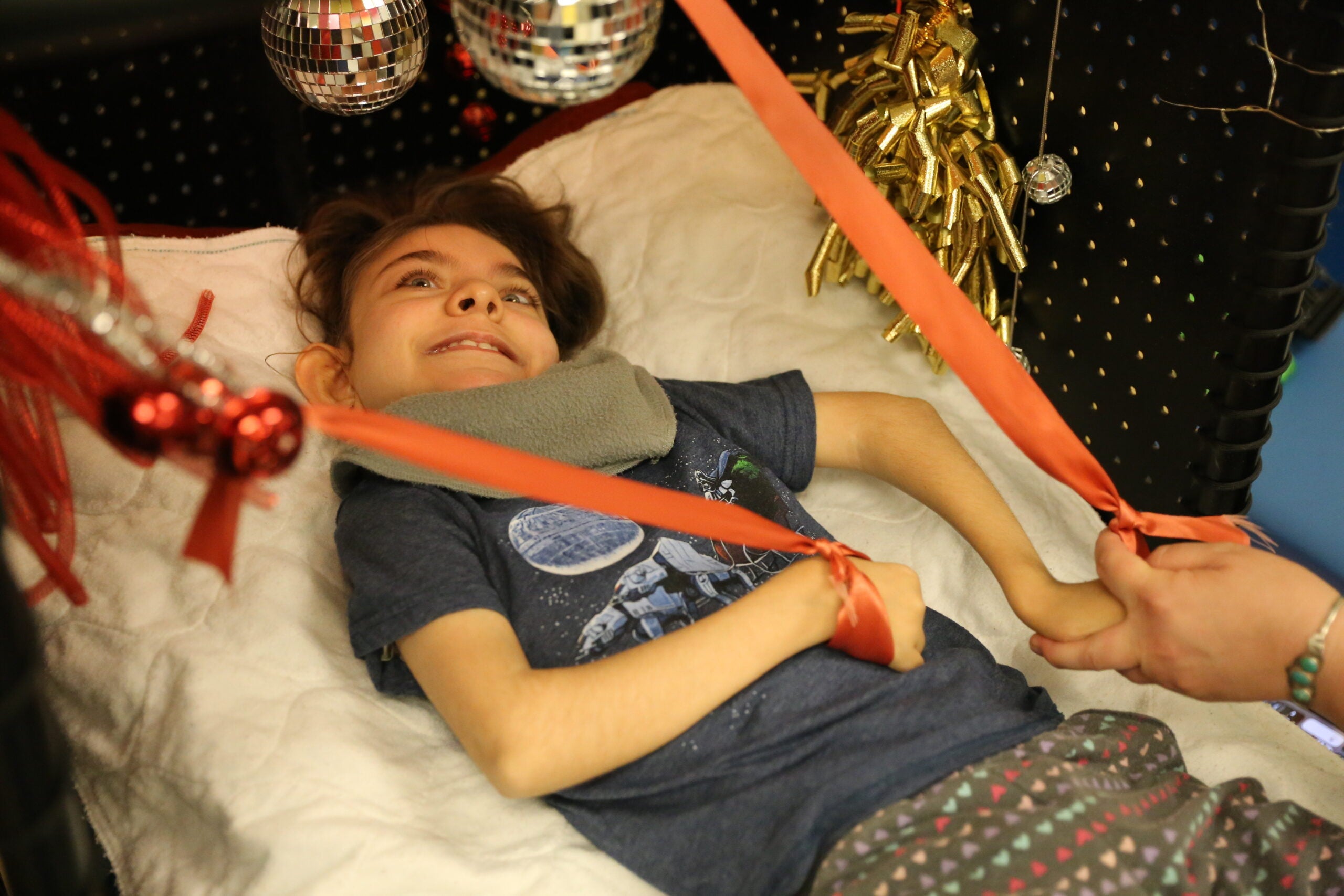 Student lays in tiny room, plays with red ribbons and smiles while looking at disco balls.