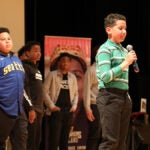 A student with a microphone stands on a stage during an event, several students stand behind the student on the stage.