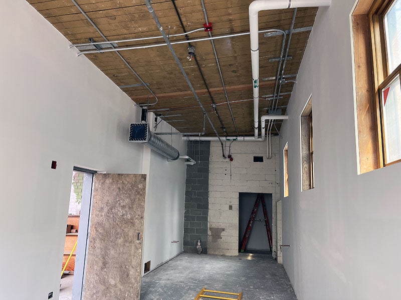 a room with piping at the ceiling, windows on the right side wall, and a door on the left side. the floor is concrete and the back wall is concrete masonry units