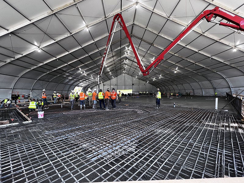 a grid of rebar is at the bottom, a group of workers in hardhats and safety vests stands in the center, a concrete pump is in the back with a large tent structure over it all