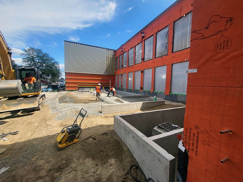 a courtyard area has soil, gravel, and concrete walls. workers are moving materials. a compactor for gravel is in the front