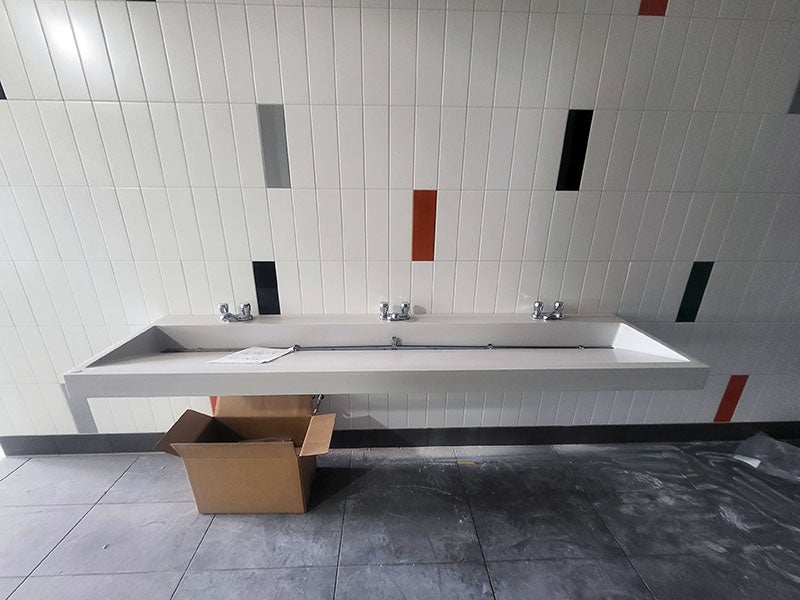 a trough sink with three faucets hangs on a tile wall with a few colored accent tiles