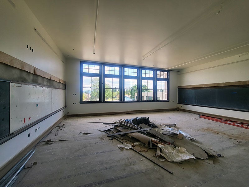 a classroom has white boards on the left wall, windows on the far wall, and a blackboard on the right wall. the floor is covered with construction protection and a pile of material is in the middle of the floor