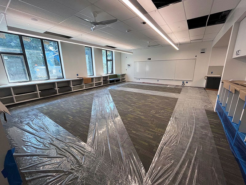 a classroom has shelves under the windows, carpet with plastic runners, and a white board