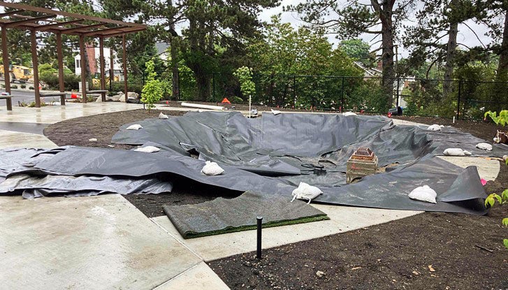 a depression surrounded by a concrete walkway has black material spread across it - a trellis-type structure and trees are in the background