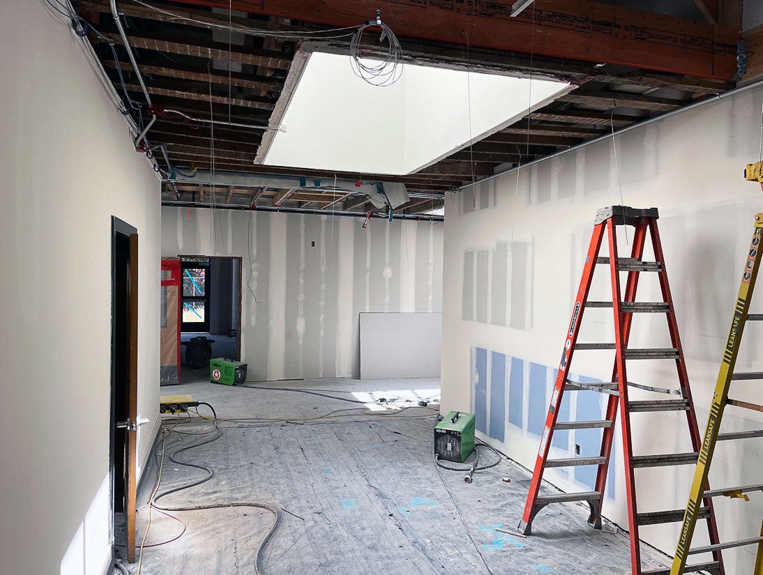 a hallway has wallboard installed and taped, an open ceiling with a skylight, and covered floors. Two ladders are in the lower right and power cords are along the floor.