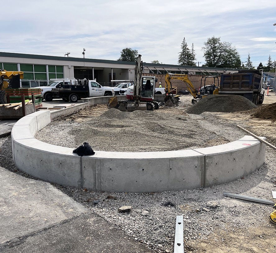 a semi-circular low wall has gravel piled in the center; construction equipment and a building are behind it