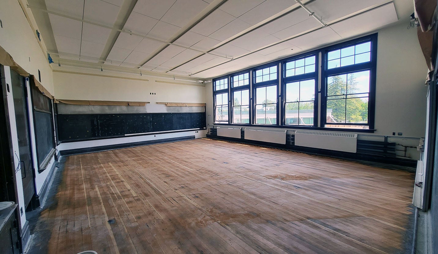 an empty room has blackboards on one wall, windows on another wall with radiators below them, and a wood floor