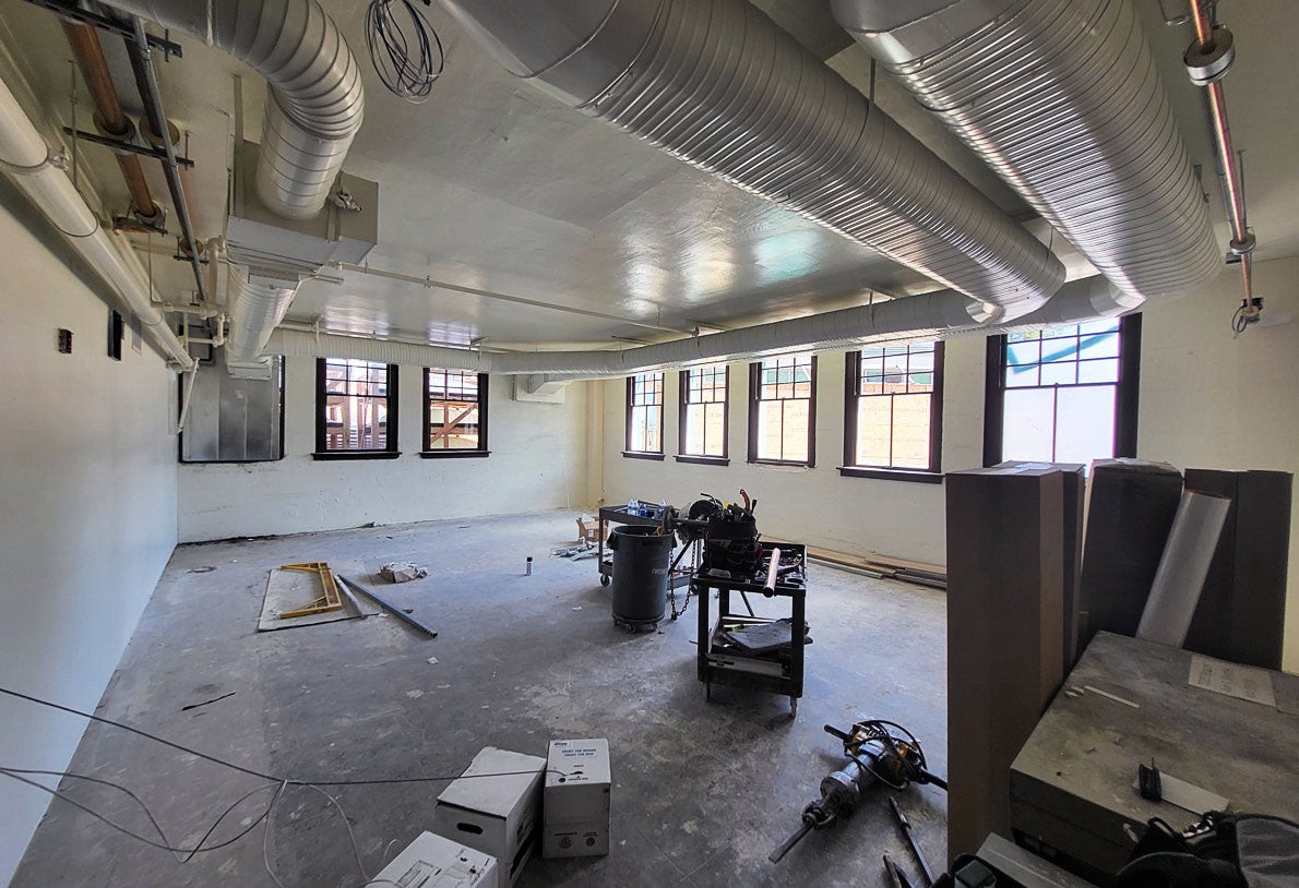 a room with windows on two walls has duct work in the ceiling. some equipment and construction tools are on the concrete floor