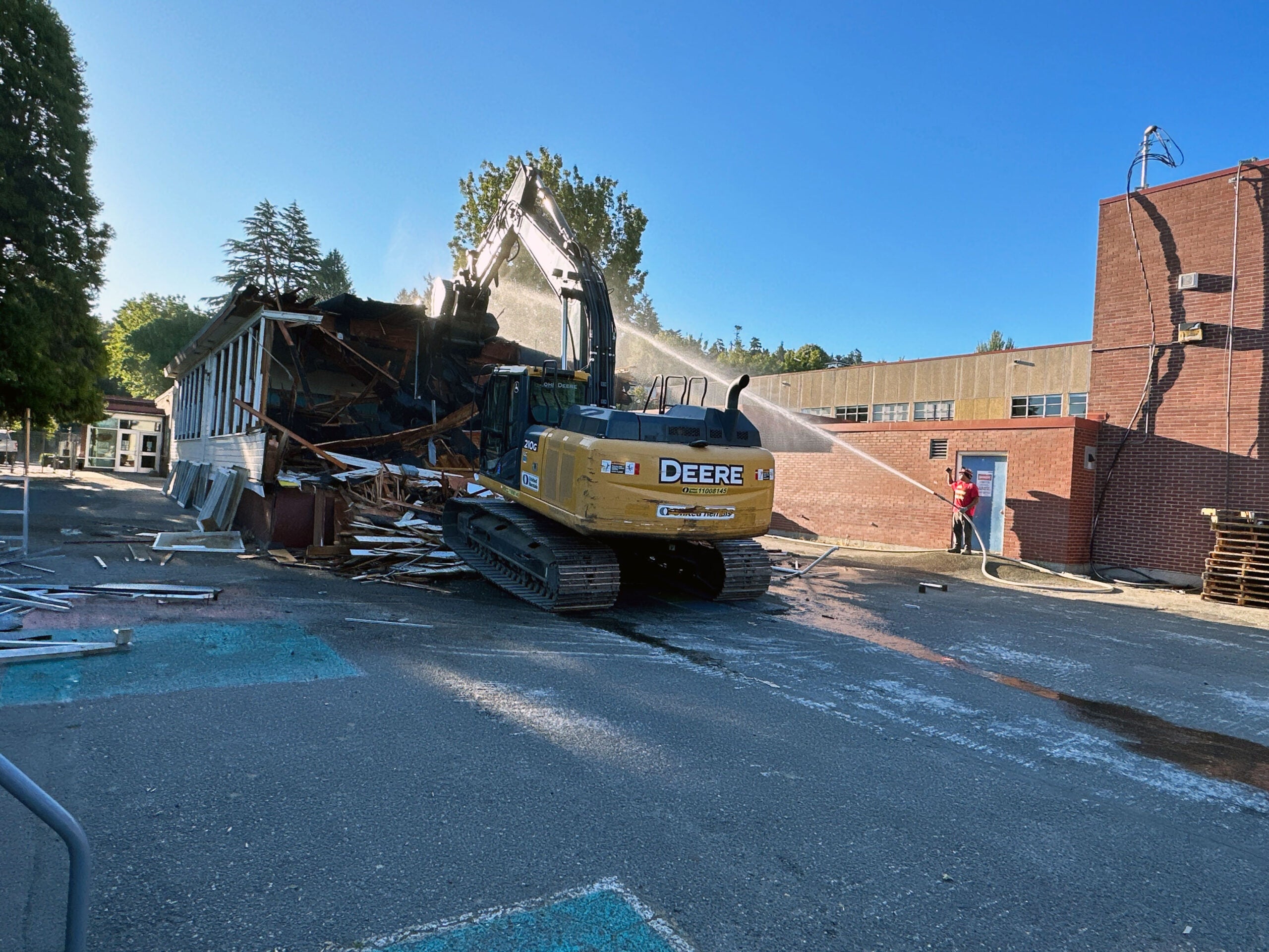 an excavator is crushing a wooden building while a person sprays a hose at it