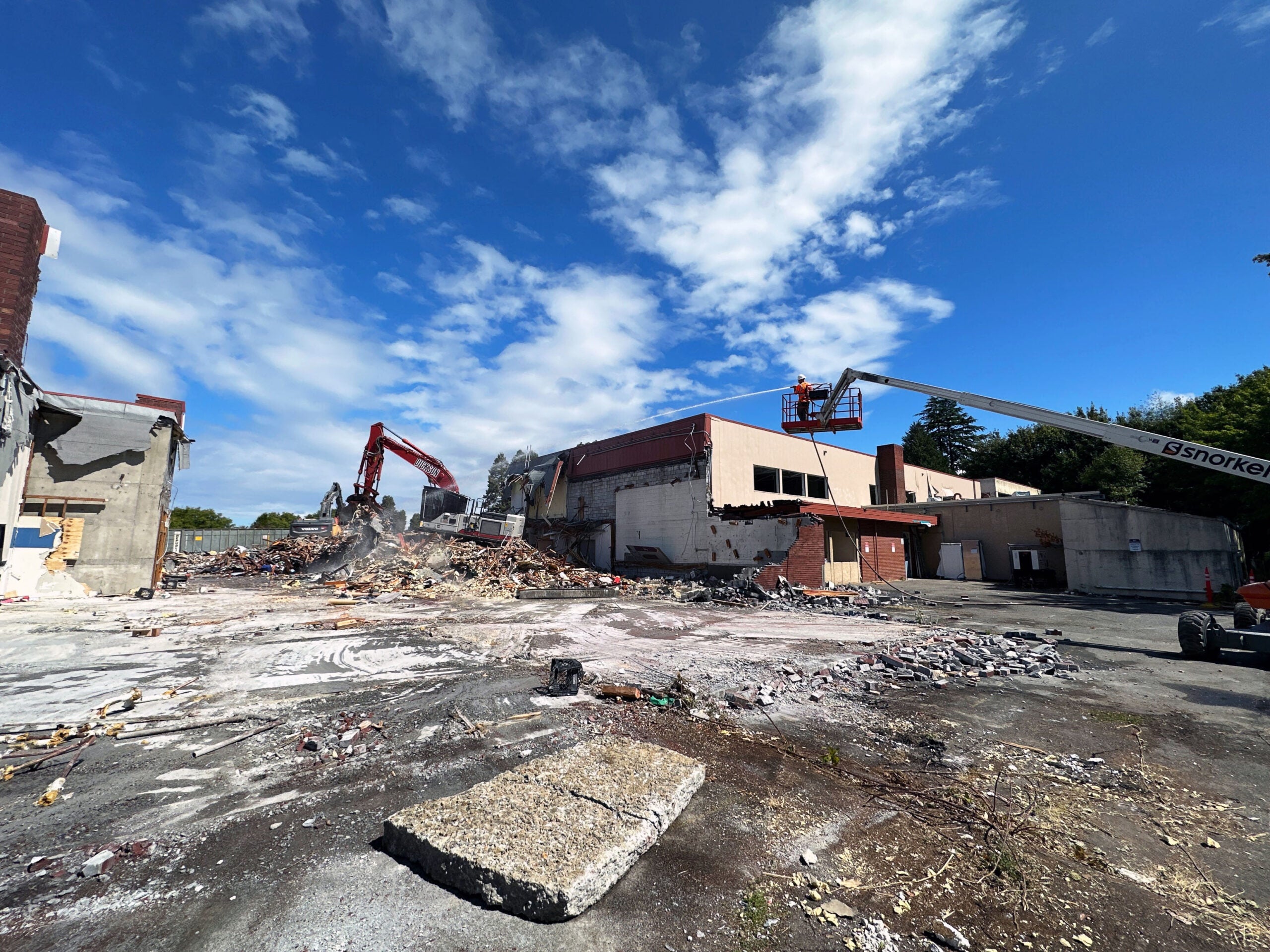 an excavator is crushing a brick building. There is a person on a lift spraying a hose. There is rubble on the concrete in the foreground