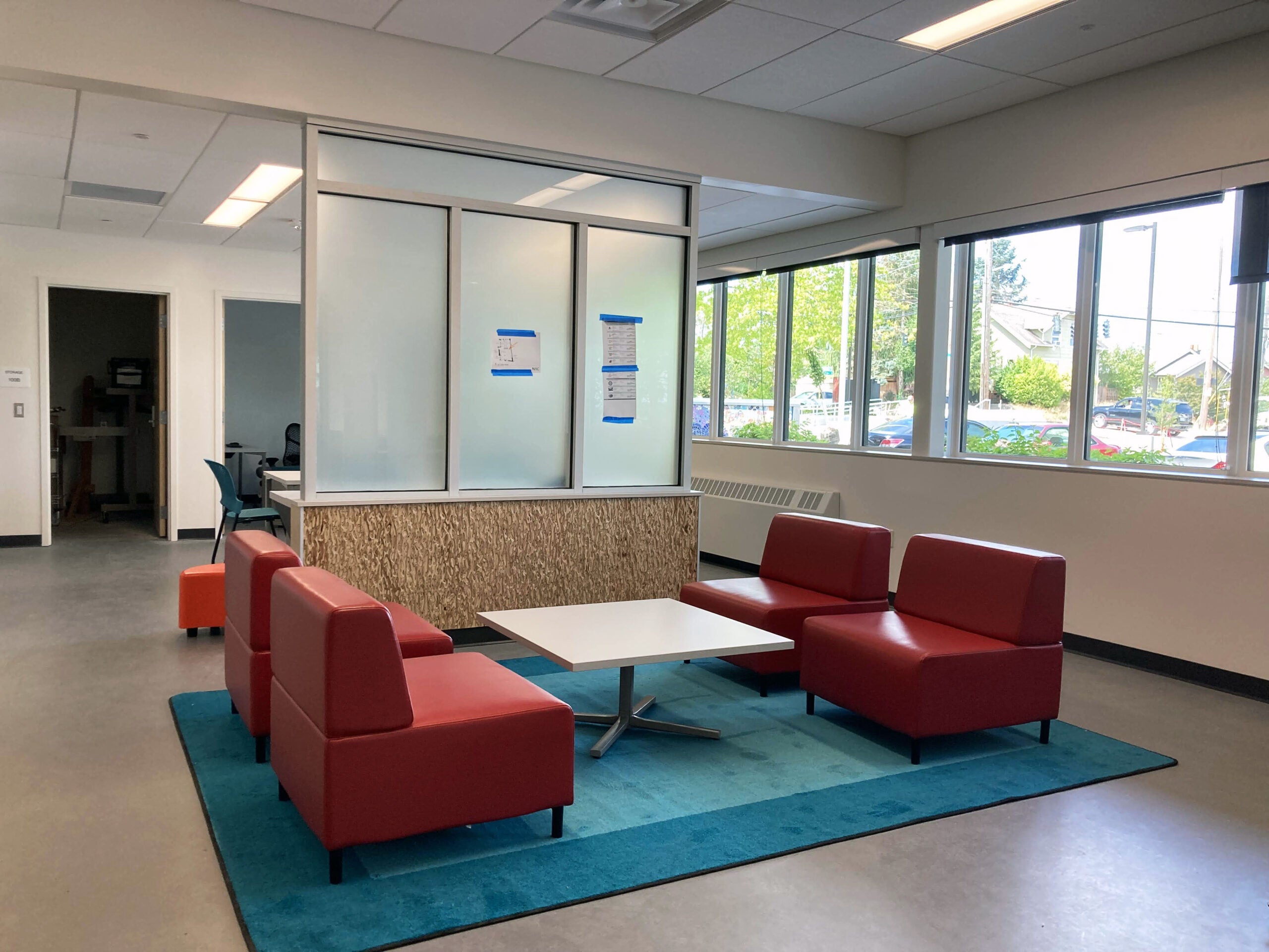 four red chairs sit on a blue area rug with a table between them. a partial wall separates them from another area. windows are on one wall